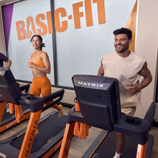 Une femme et un homme s&rsquo;entra&icirc;nent c&ocirc;te &agrave; c&ocirc;te sur des tapis de course chez Basic-Fit, tout en souriant. Ils courent dans une salle lumineuse avec une grande enseigne Basic-Fit visible en arri&egrave;re-plan.